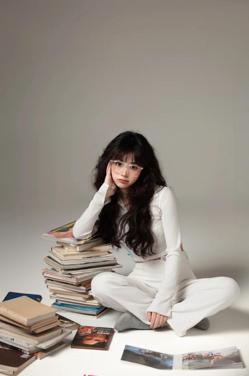 (ngồi bệt, tay chống cằm, tựa vào chồng sách)