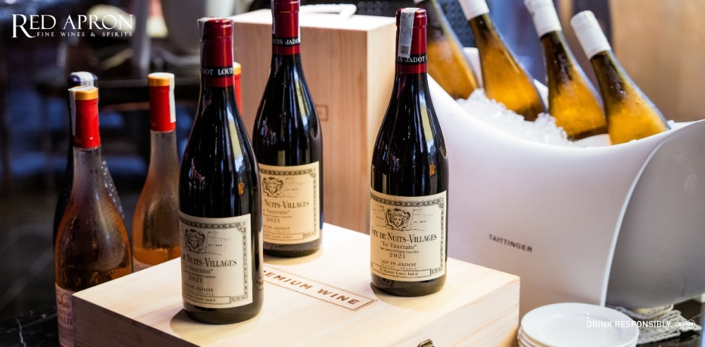 Nhiều chai rượu vang Maison Louis Jadot đặt trong thùng gỗ và xô đá trắng, minh họa nội dung về nhập khẩu chính ngạch, bảo quản kho lạnh và trải nghiệm mua rượu vang tại Red Apron.