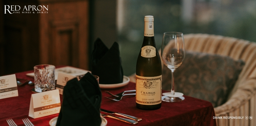 Một chai rượu vang Louis Jadot Beaujolais-Villages trên khăn trải bàn đỏ cùng ly rượu và nến trang trí, thể hiện phong cách vang đỏ tươi mát của vùng Beaujolais mà bài viết mô tả.