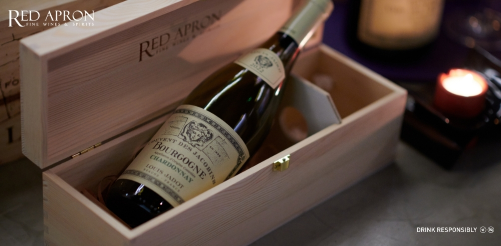 Cận cảnh chai rượu vang trắng Louis Jadot Bourgogne Chardonnay nằm trong hộp gỗ in logo Red Apron, phù hợp nội dung về vang trắng Burgundy thanh mảnh cho hải sản và salad.