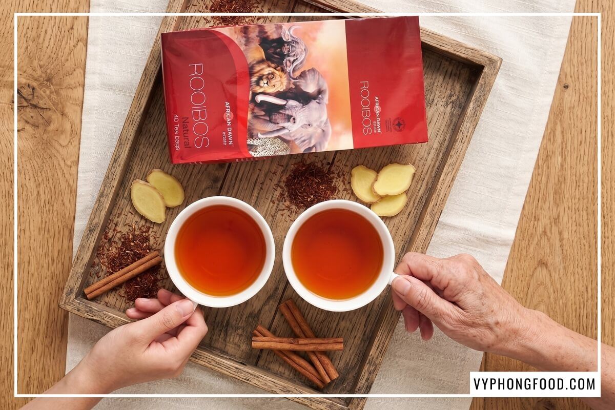 Khay gỗ sồi bày hộp trà rooibos Nam Phi thương hiệu African Dawn hai tách trà rooibos đỏ hổ phách cùng vài lát gừng và thanh quế.