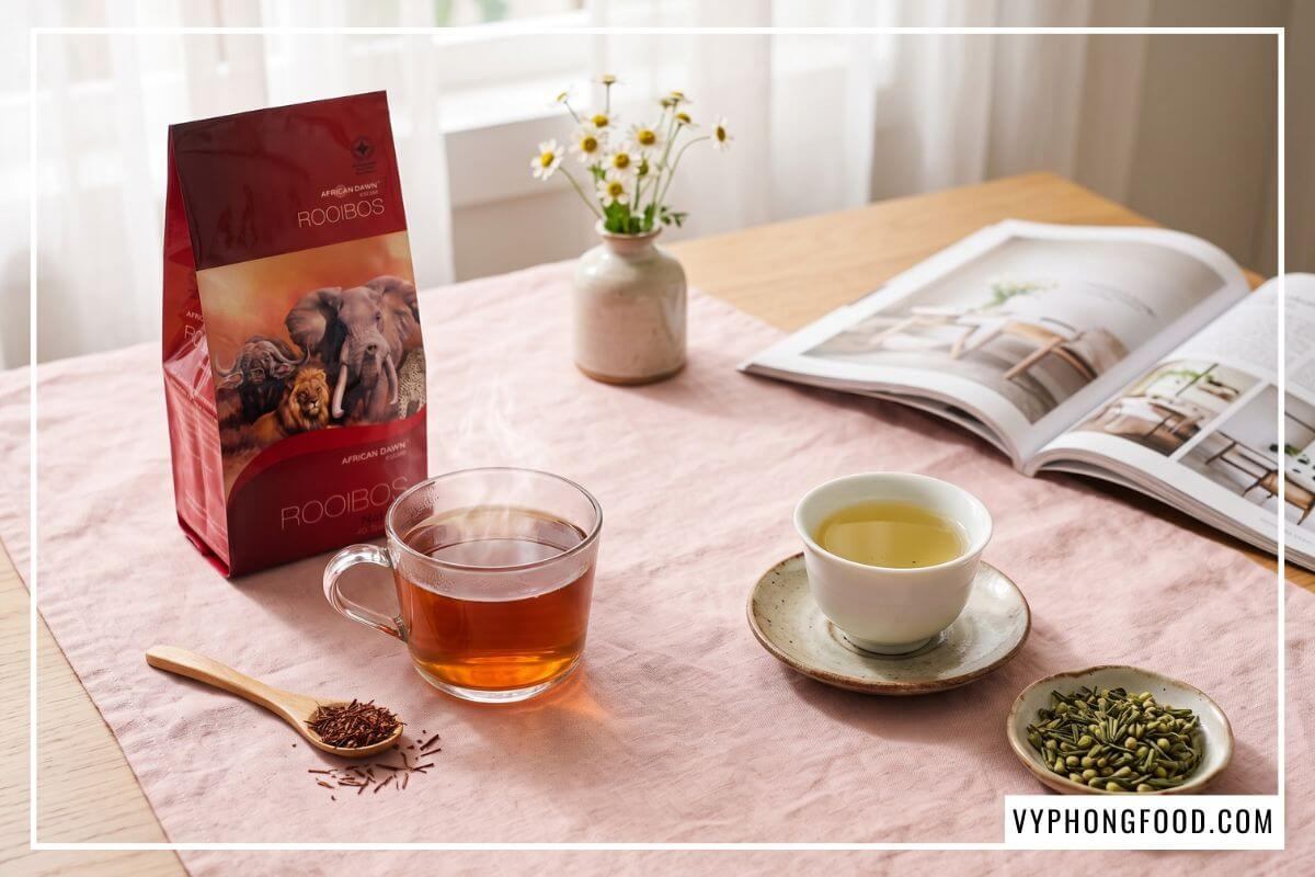 So sánh hồng trà rooibos và trà tâm sen trị mất ngủ đặt trên bàn trải khăn hồng pastel êm dịu.