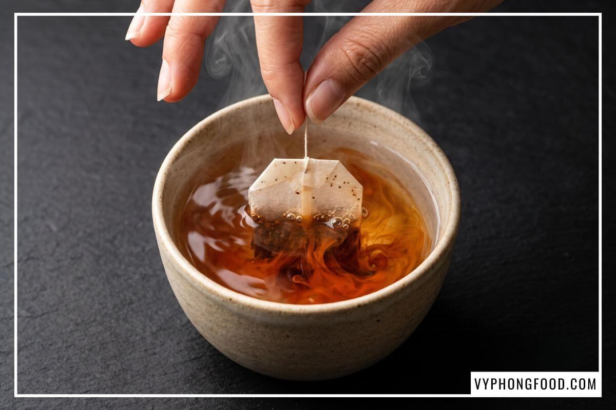 Túi lọc trà thảo mộc rooibos không caffeine bung tỏa sắc đỏ nâu hổ phách trong tách nước sôi trên mặt đá phiến đen.
