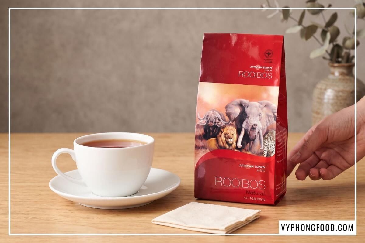 Gói hồng trà rooibos Nam Phi dạng túi lọc đứng cạnh tách trà sứ màu đỏ đất
