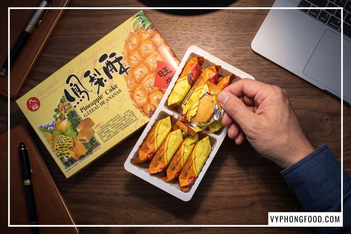 Tay nam giới xé túi nilon lấy chiếc bánh dứa nướng từ hộp Nice Choice 227g trên bàn làm việc gỗ óc chó.