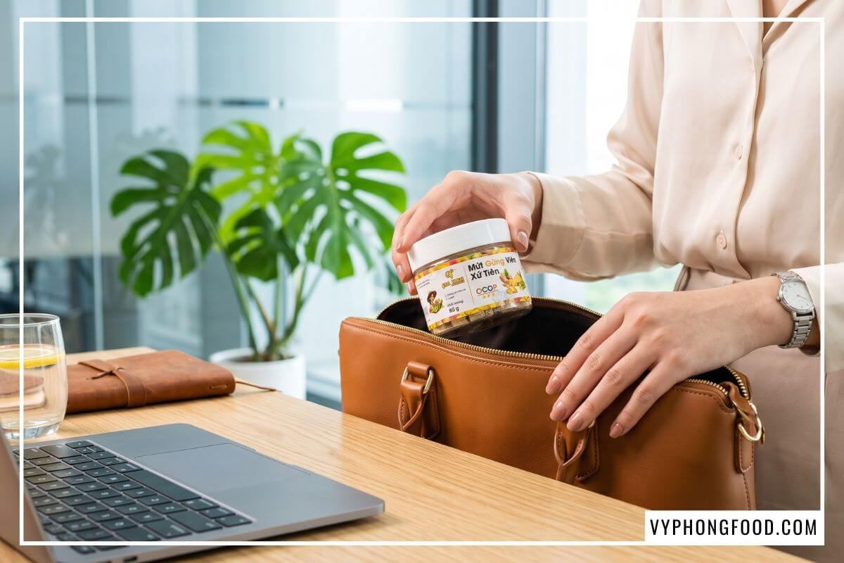Hũ mứt gừng viên 95g nắp trắng nhô ra từ miệng túi xách da đặt trên bàn làm việc gỗ sồi sáng màu.