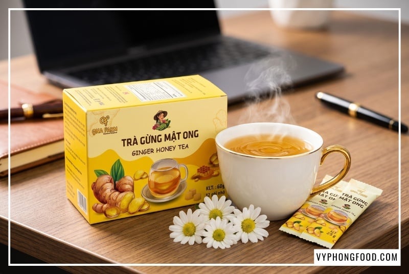 Hộp sản phẩm Trà gừng mật ong hòa tan QNA Farm 60g cao cấp đặt cạnh ly trà nóng bốc khói trên bàn làm việc văn phòng, minh họa cho công thức trà gừng giữ ấm và bảo vệ sức khỏe dân công sở tại VyPhongFood.