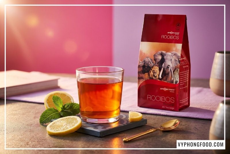 Ly trà đỏ Nam Phi pha chế sóng sánh màu hổ phách bên cạnh gói trà túi lọc Rooibos African Dawn trên bàn trà gia đình Việt do Vy Phong Food cung cấp.