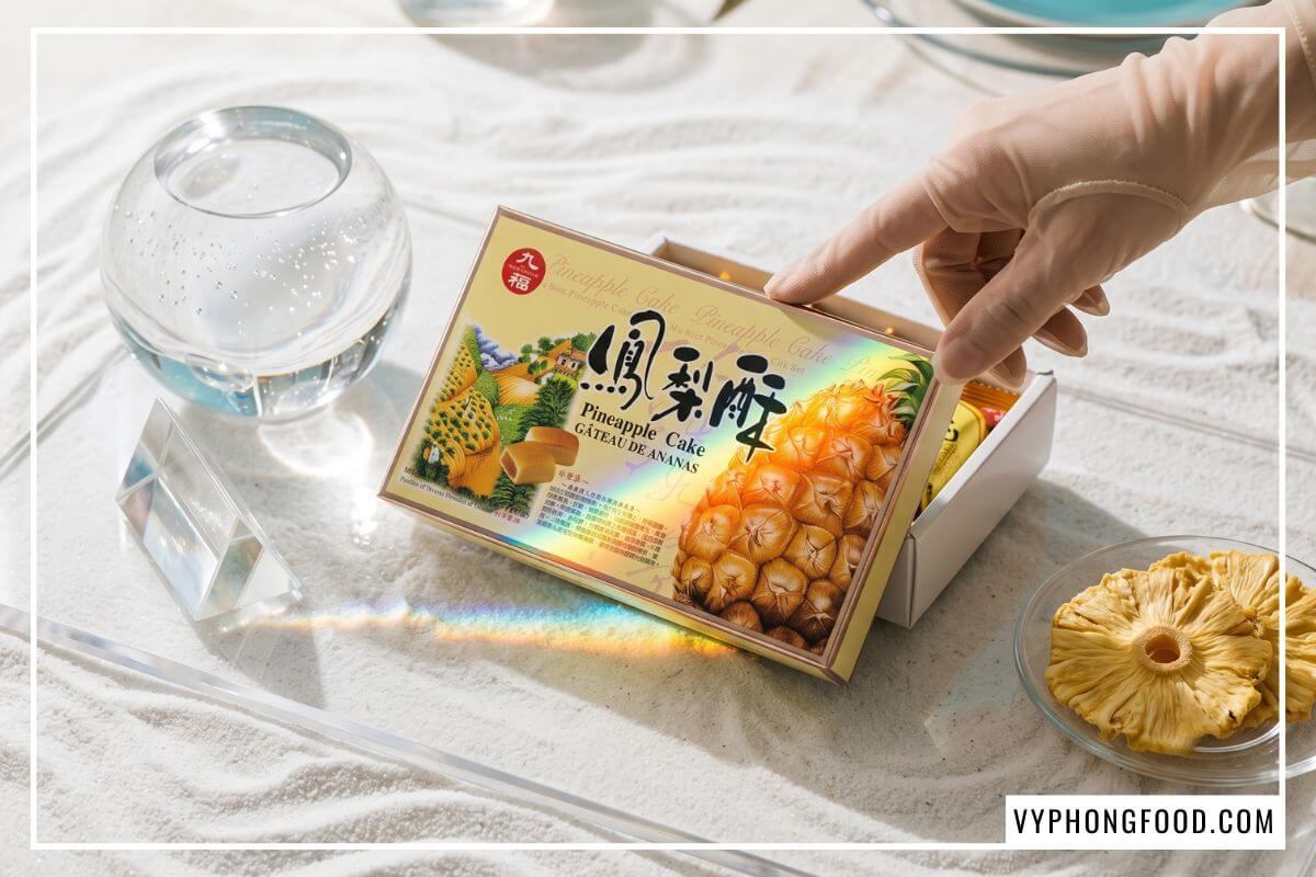 Bàn tay chạm nhẹ hộp đựng bánh dứa Đài Loan mở nắp dưới vệt sáng cầu vồng bên lát dứa sấy dẻo.