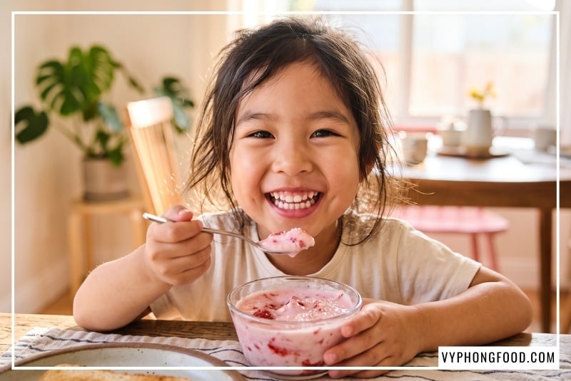 Bé gái đang ăn sữa chua dâu tây rắc đường cỏ ngọt hữu cơ không sinh axit gây sâu răng. Bài viết giải đáp chi tiết tác dụng của đường ăn kiêng đối với sức khỏe trẻ nhỏ và gợi ý địa chỉ mua đường cỏ ngọt stevia ở đâu an toàn, chính hãng.