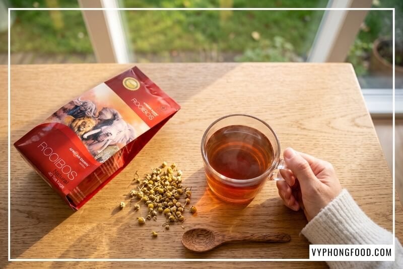 Tách hồng trà rooibos Nam Phi dạng túi lọc màu đỏ hổ phách ấm áp trên bàn gỗ sồi rợp nắng.