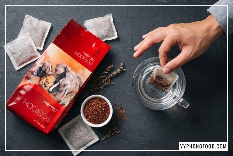 Bàn tay thả túi lọc hồng trà rooibos Nam Phi vào chén nước nóng trên nền đá ghi xám.