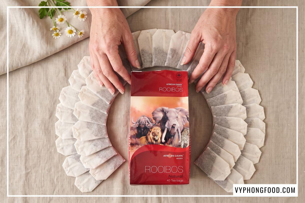 Nằm bao quanh gói hồng trà rooibos Nam Phi thương hiệu African Dawn cắt phần đầu là 40 gói trà túi lọc trắng ngà xếp hình rẽ quạt, nổi bật trên nền vải be phẳng phiu có điểm xuyết vài nhành hoa cúc li ti như một liệu pháp trà thảo mộc giảm căng thẳng.