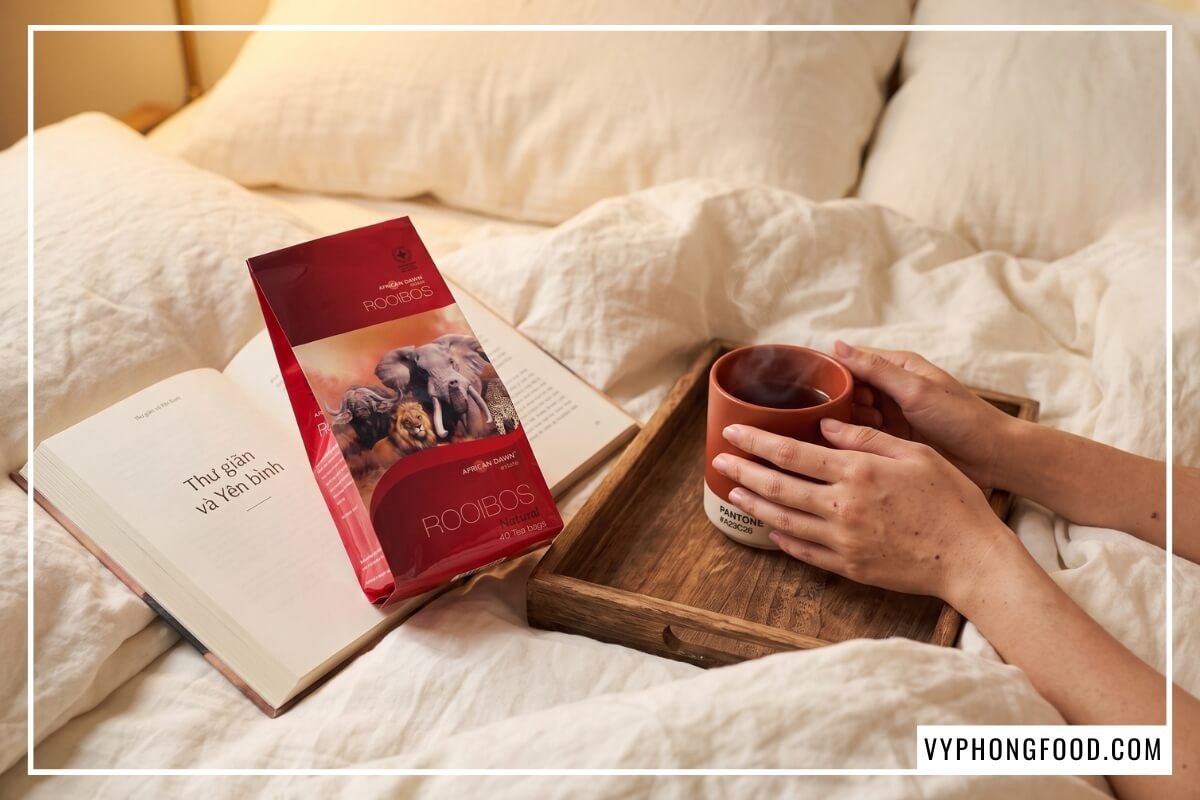 Bàn tay ôm ly trà ấm bên gói hồng trà rooibos Nam Phi African Dawn trên giường, xóa tan âu lo trà rooibos có gây mất ngủ không lúc nửa đêm.