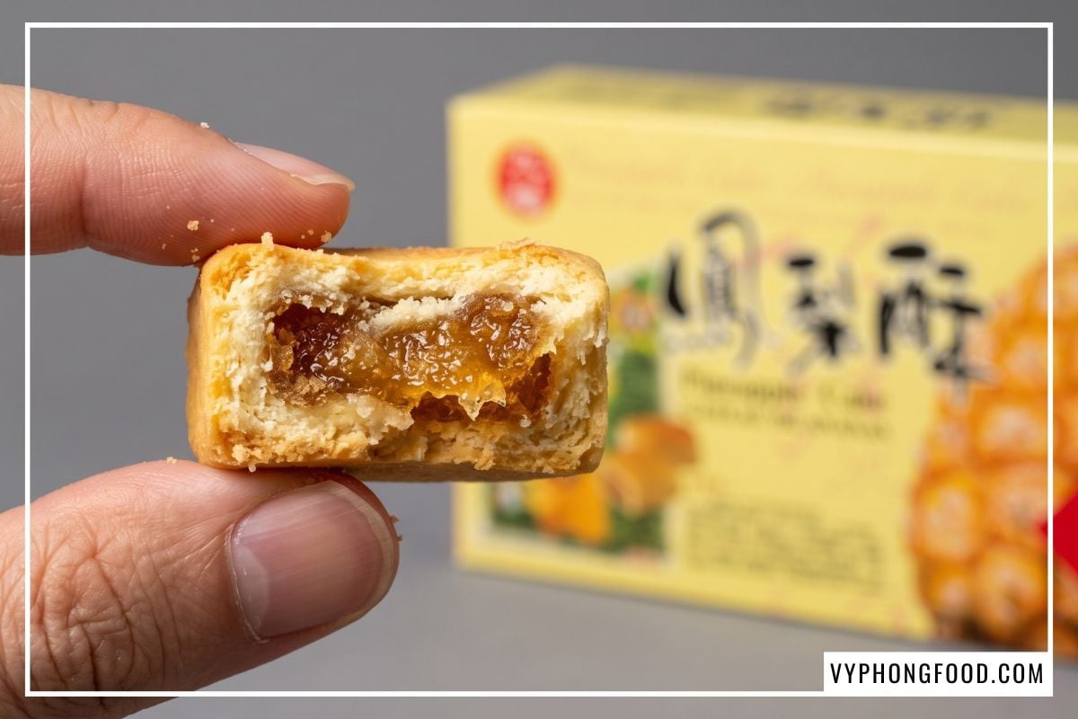 Cận cảnh chiếc bánh dứa cắn dở lòi lõi mứt óng ả khoe vỏ xốp mềm, phía sau là hộp Pineapple Cake Taiwan Nice Choice mờ ảo.