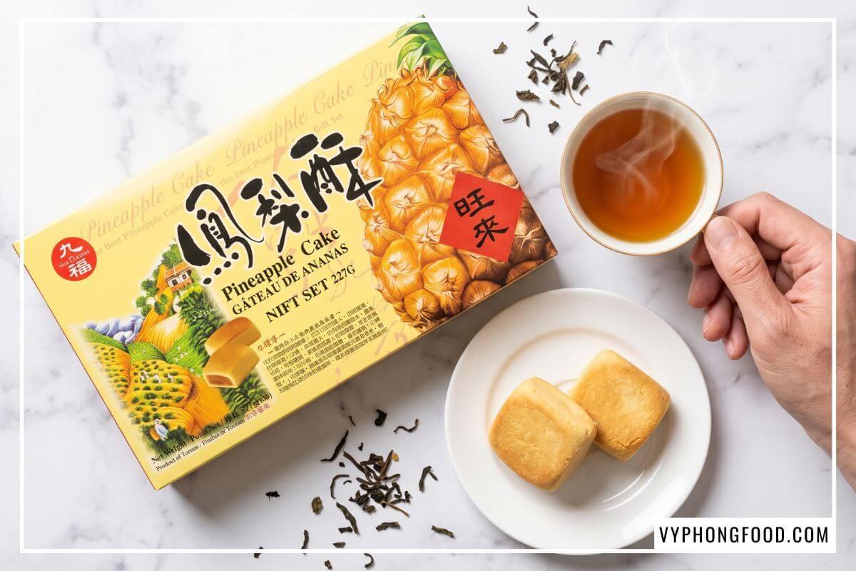 Hộp bánh dứa Đài Loan Nice Choice màu vàng tươi nằm cạnh chén trà Ô Long đang bốc khói trên bàn đá hoa cương.