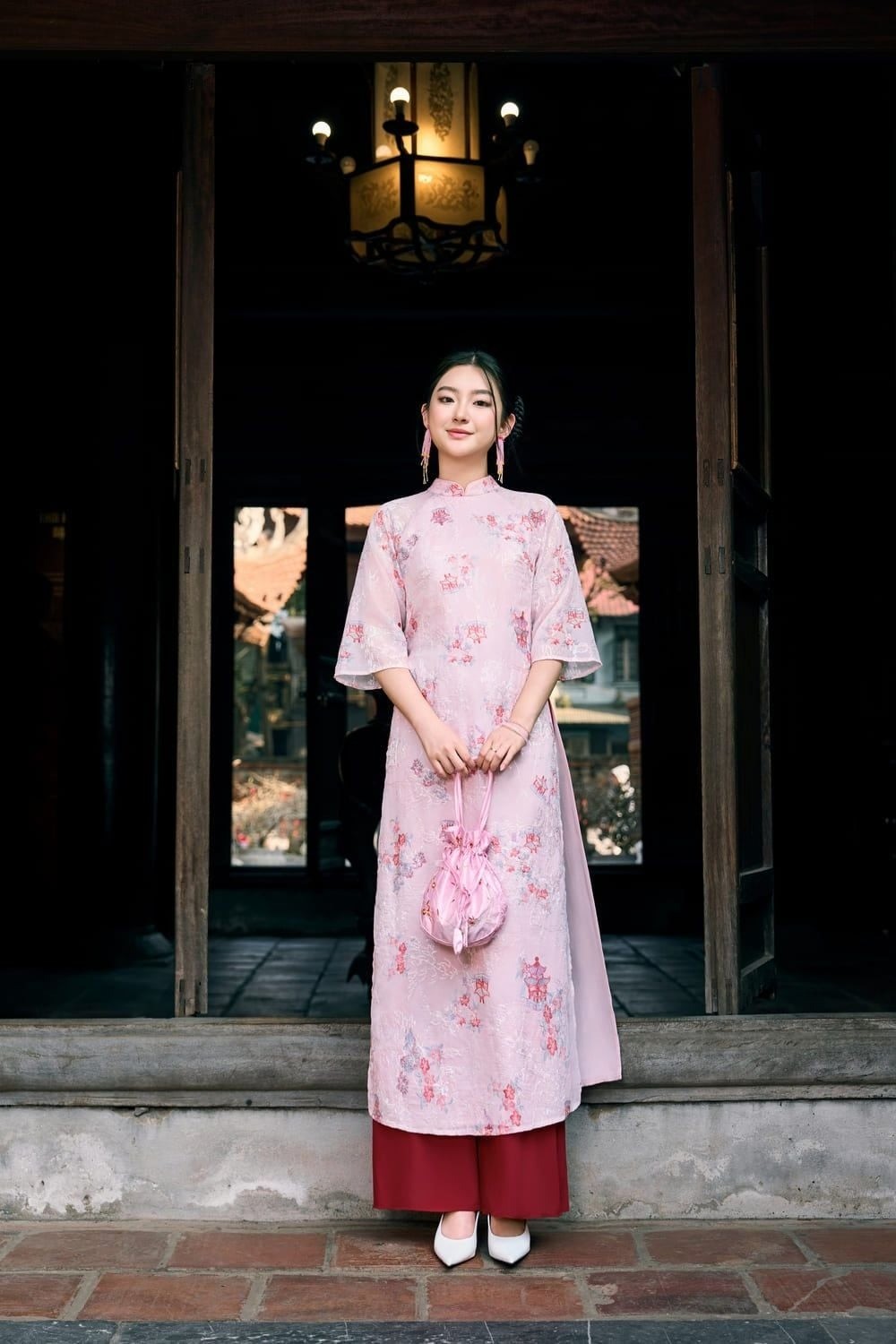 Đứng giữa khung cửa, tay xách túi nhỏ, mắt nhìn trực diện, góc chụp toàn thân từ xa