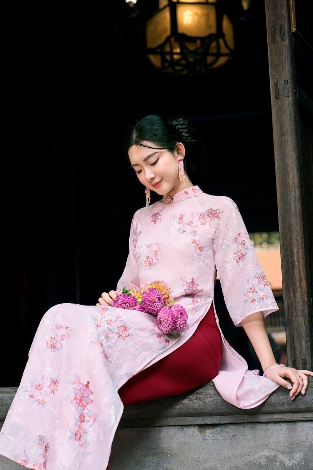 Ngồi trên bậu cửa, mắt nhìn xuống bó hoa, biểu cảm dịu hiền, góc chụp trung cảnh