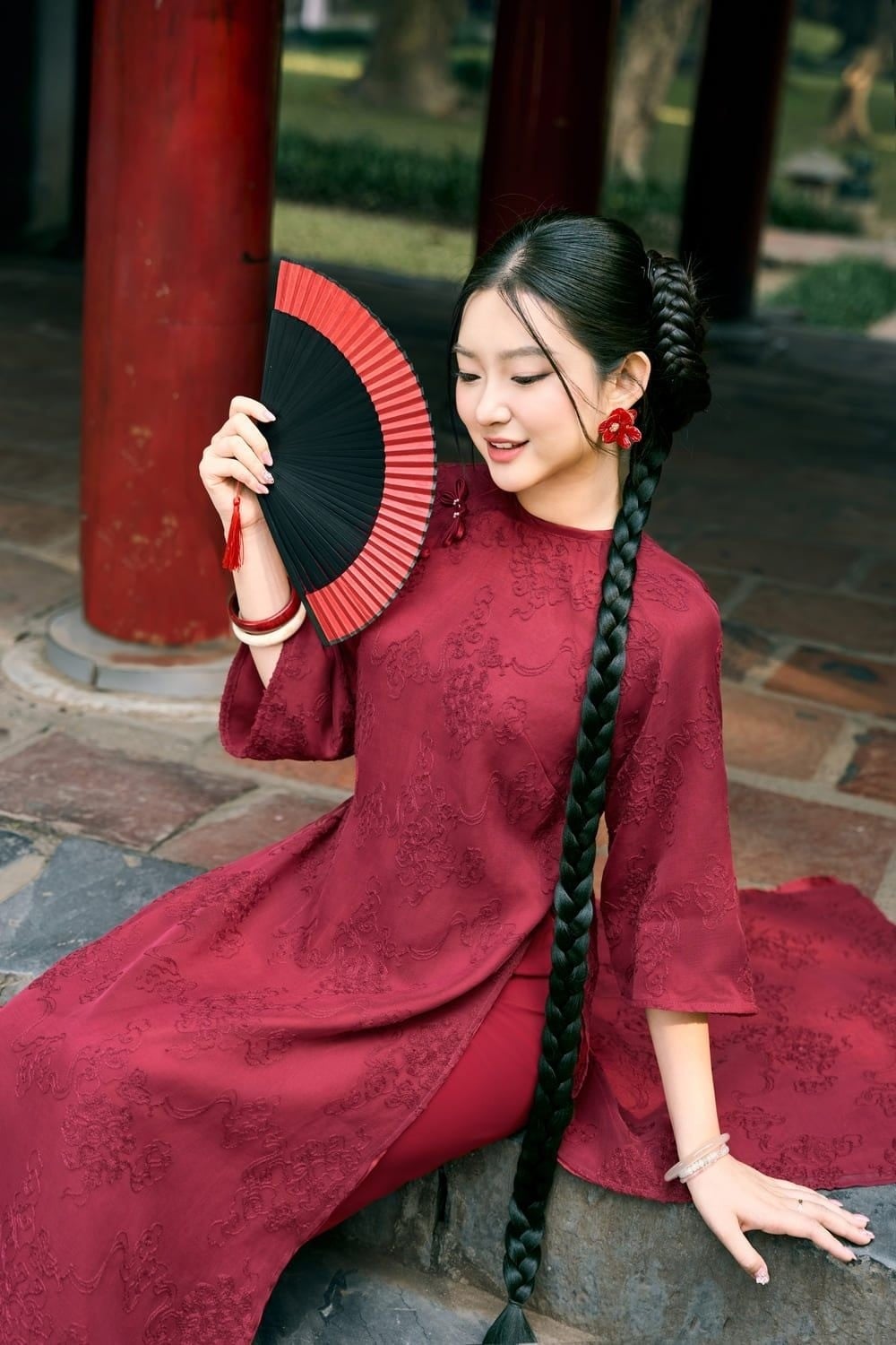 Ngồi xếp bằng, tay cầm quạt đỏ đen mở rộng, mắt nhìn xuống quạt, góc chụp trung cảnh