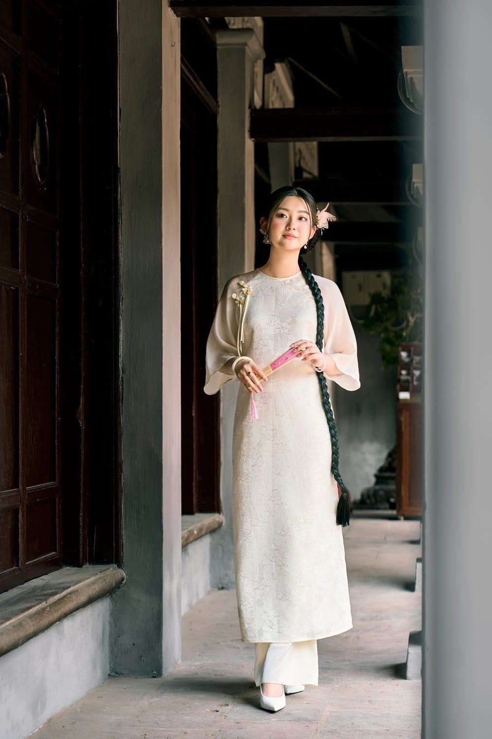 Bước đi trong hành lang, tay cầm quạt hồng nhỏ, mắt nhìn phía trước, góc chụp toàn thân