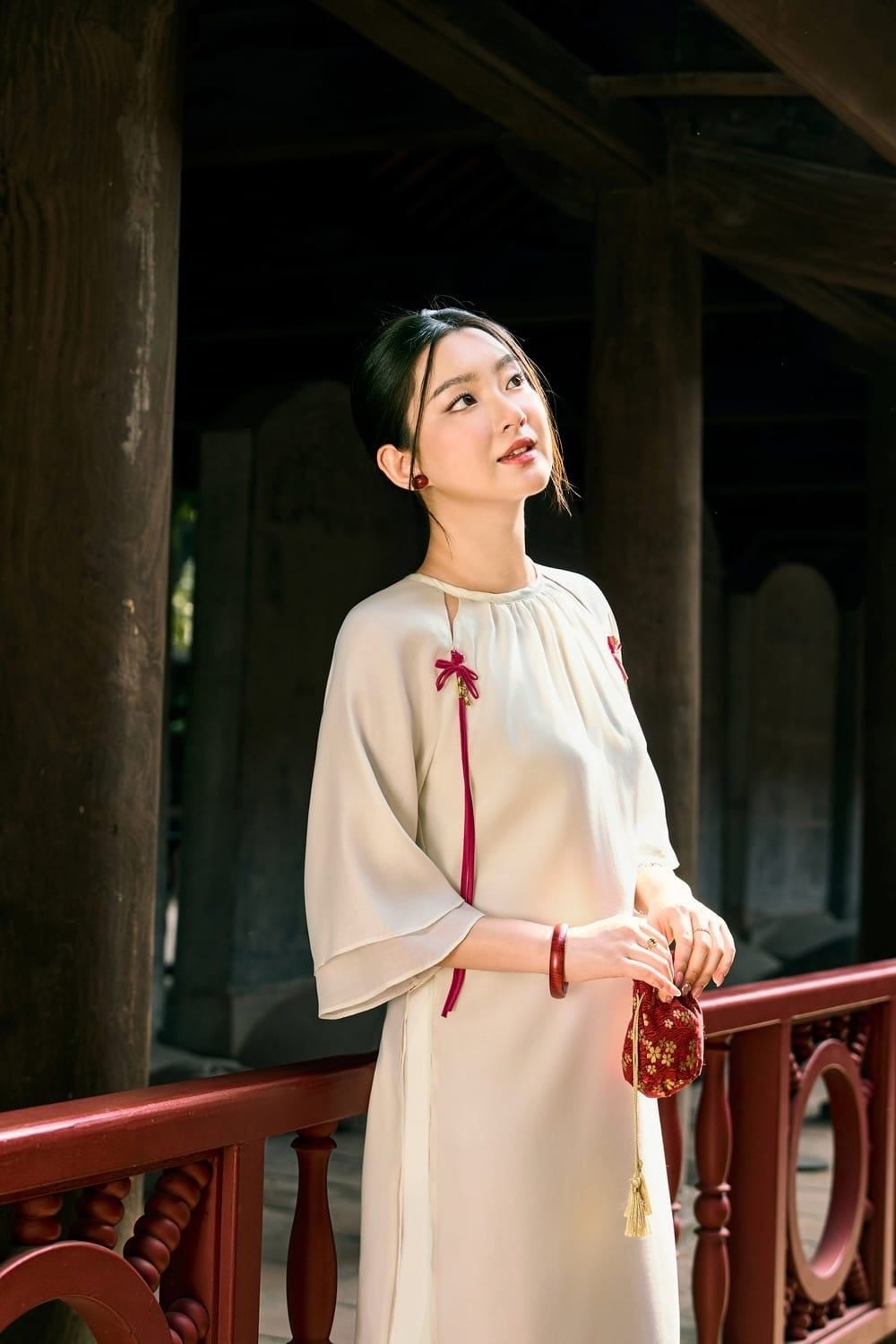 Tựa tay vào lan can, tay cầm túi lụa đỏ, mắt nhìn hướng lên, góc chụp cận trung