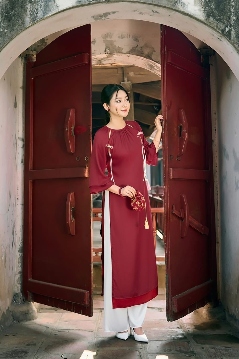 Đứng giữa lối đi cổng tròn, tay cầm túi nhỏ, mắt nhìn sang bên, góc chụp toàn thân