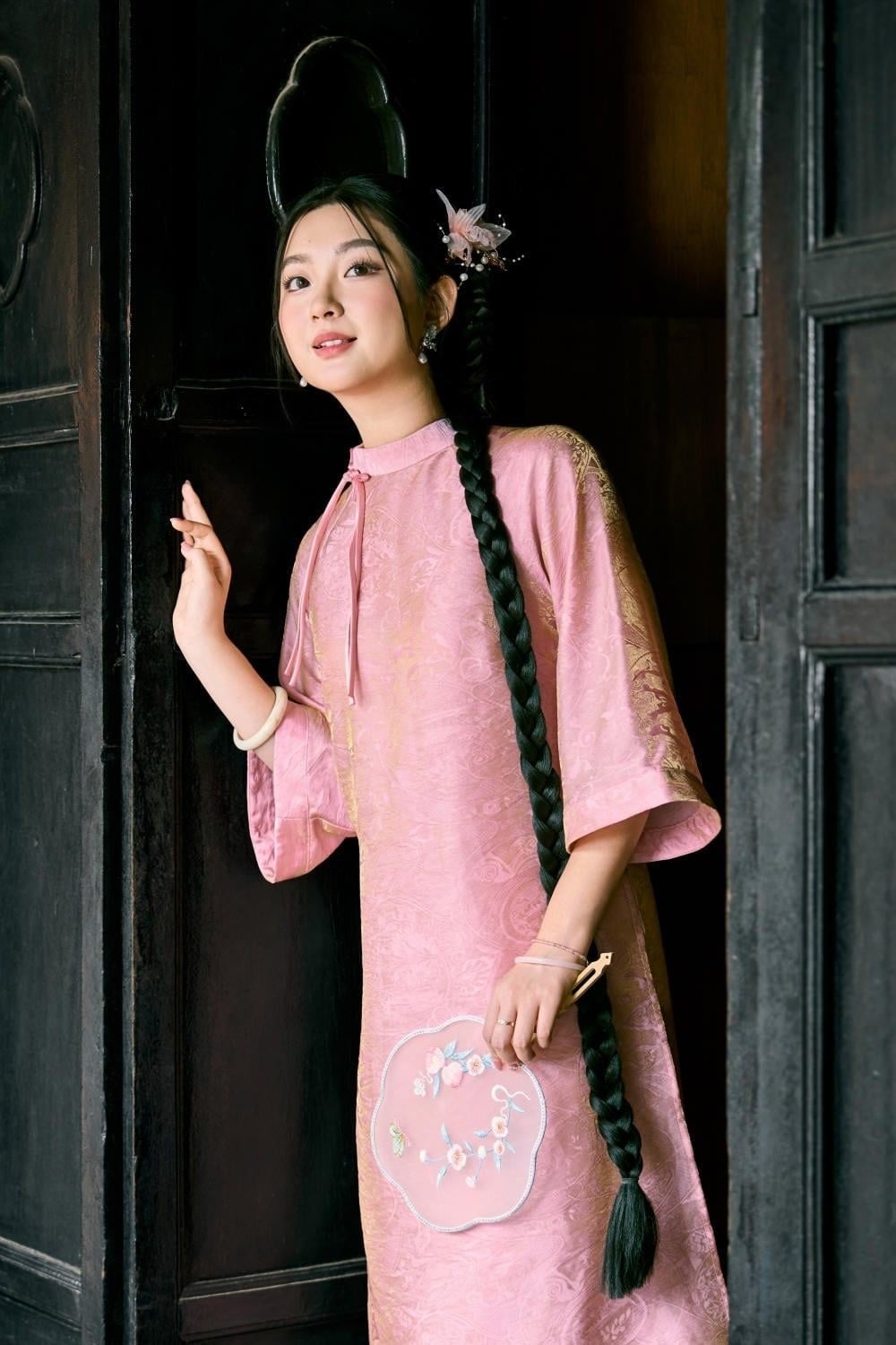 Tựa lưng vào cửa gỗ, tay cầm quạt tròn, mắt nhìn lên cao, góc chụp cận trung