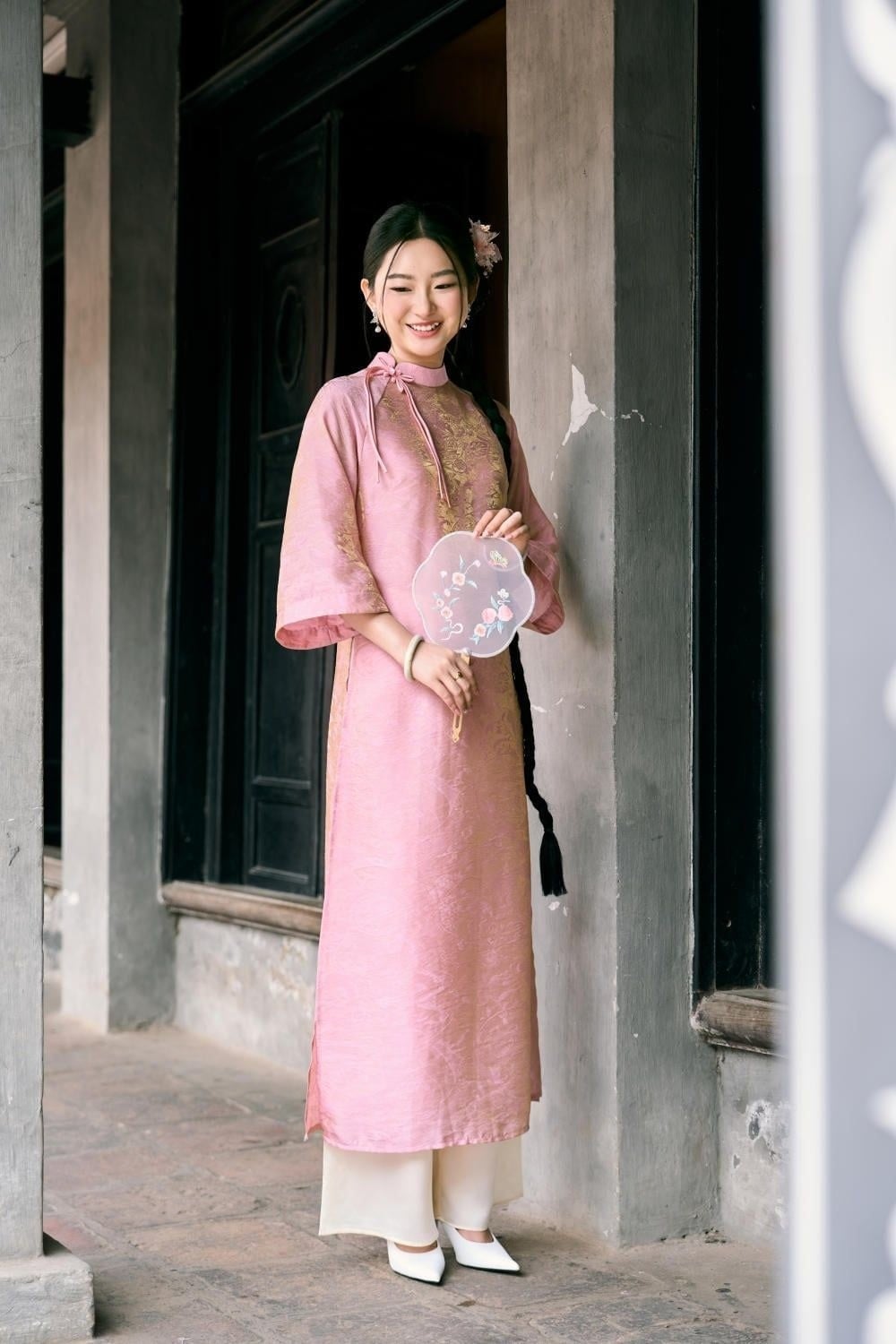 Đứng chính diện cửa, tay cầm quạt tròn bằng hai tay, mắt nhìn ống kính, góc chụp toàn thân