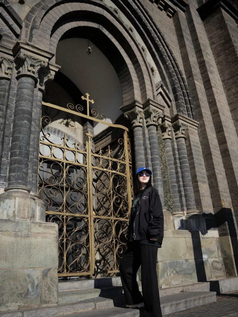 Đứng tựa cổng sắt, một tay đút túi, đeo kính râm