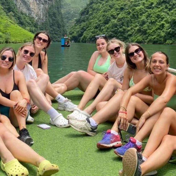 A group of young women wearing sunglasses sitting and relaxing on the green deck of a tour boat on the calm Nho Que River.