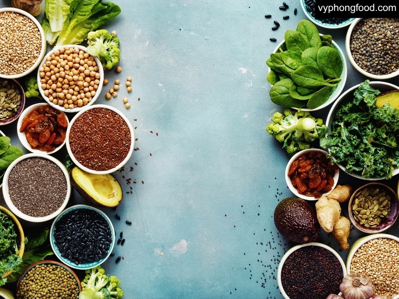 Ảnh bột ngũ cốc dinh dưỡng pha sẵn trong ly, kèm tô ngũ cốc – hạt nguyên liệu (yến mạch, đậu, mè, hạt lanh, óc chó) trên nền sáng; minh họa giải pháp đủ đạm thực vật cho người ăn chay.