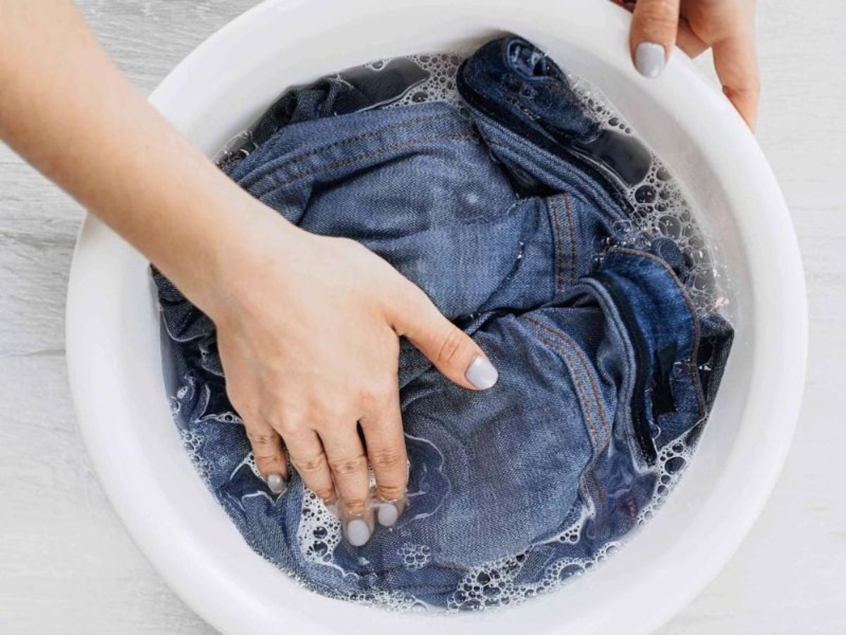 Bàn tay giặt quần jean xanh trong chậu nước xà phòng để làm sạch bề mặt vải.