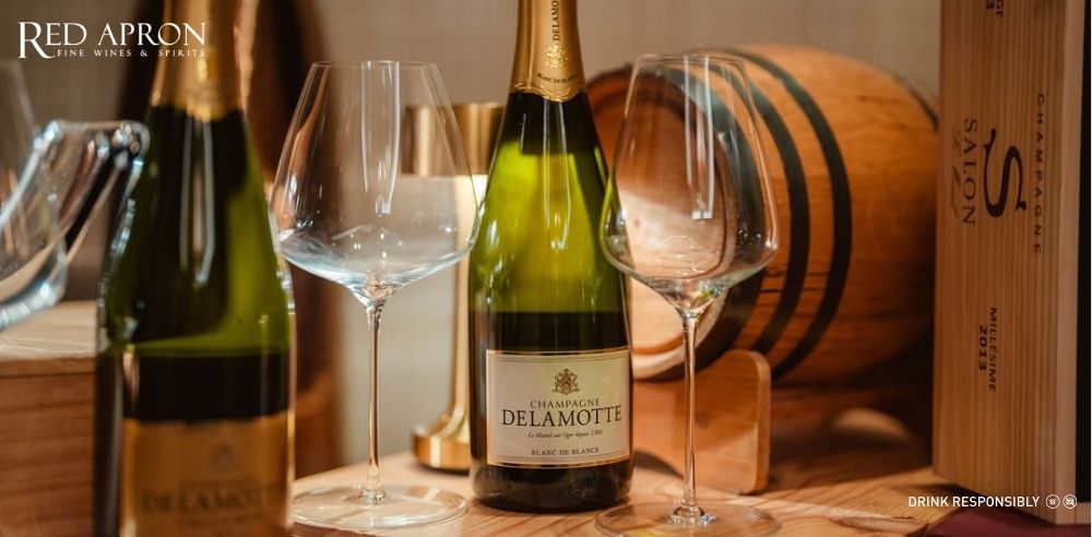 Chai Champagne Delamotte Blanc de Blancs đặt giữa hai ly tulip trong bối cảnh hầm/barel gỗ, minh họa bảo quản mát và phục vụ đúng cách; watermark Red Apron.