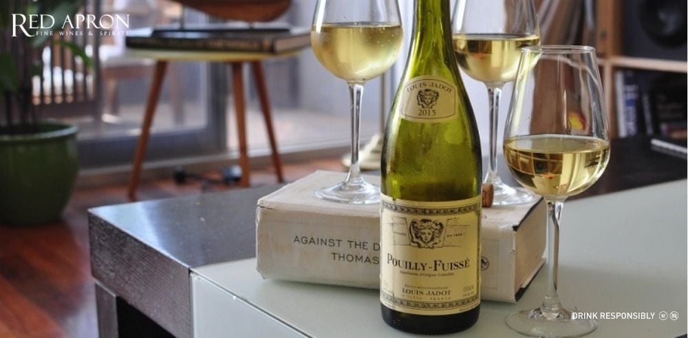 Chai rượu vang trắng Louis Jadot Pouilly-Fuissé đặt trên chồng sách cùng nhiều ly vang, trong không gian phòng khách ấm cúng.