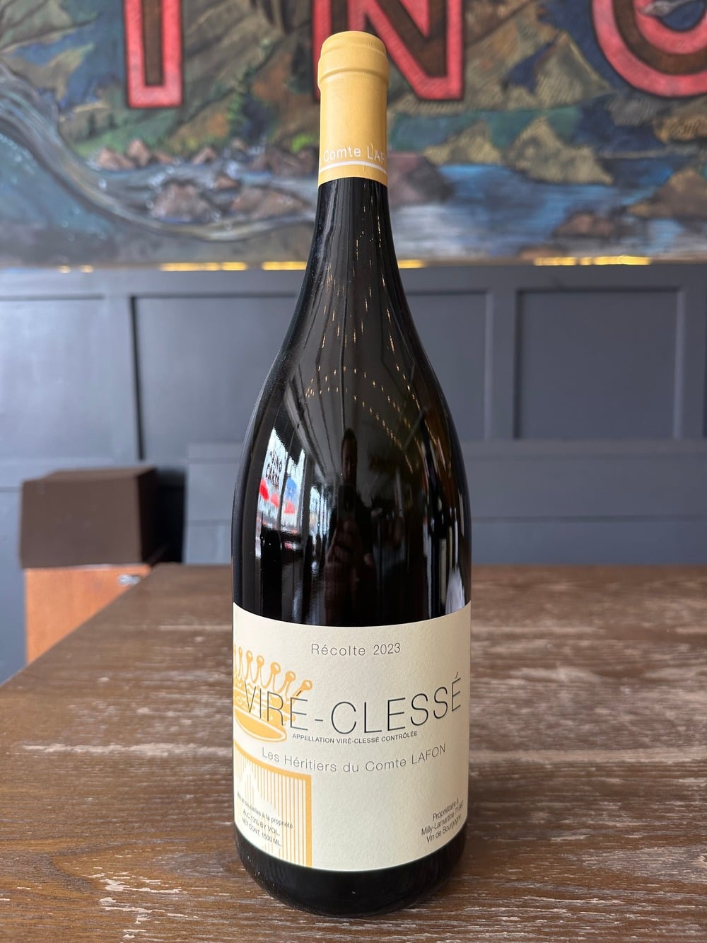 Cận cảnh chai rượu Comte Lafon Les Héritiers Viré-Clessé Récolte 2023 đặt trên mặt bàn gỗ, nền là bức tranh lớn trong quán rượu.