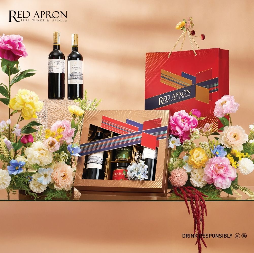 Bộ quà Tết Red Apron gồm chai rượu vang, túi và hộp quà đặt giữa hoa trang trí, gợi ý trình bày quà Tết cho sui gia