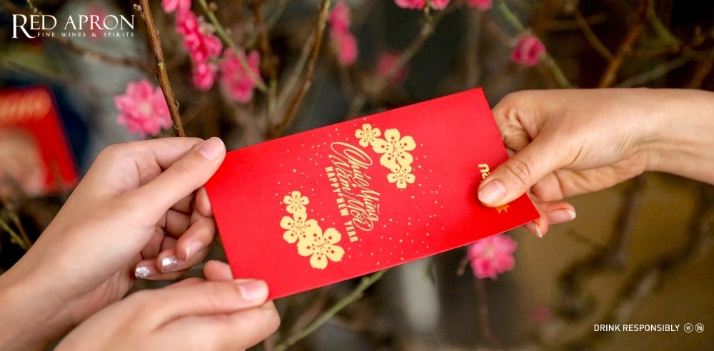 Cận cảnh hai tay trao bao lì xì đỏ có họa tiết hoa và chữ “Happy New Year”, minh họa ý tưởng bao lì xì handmade dịp Tết