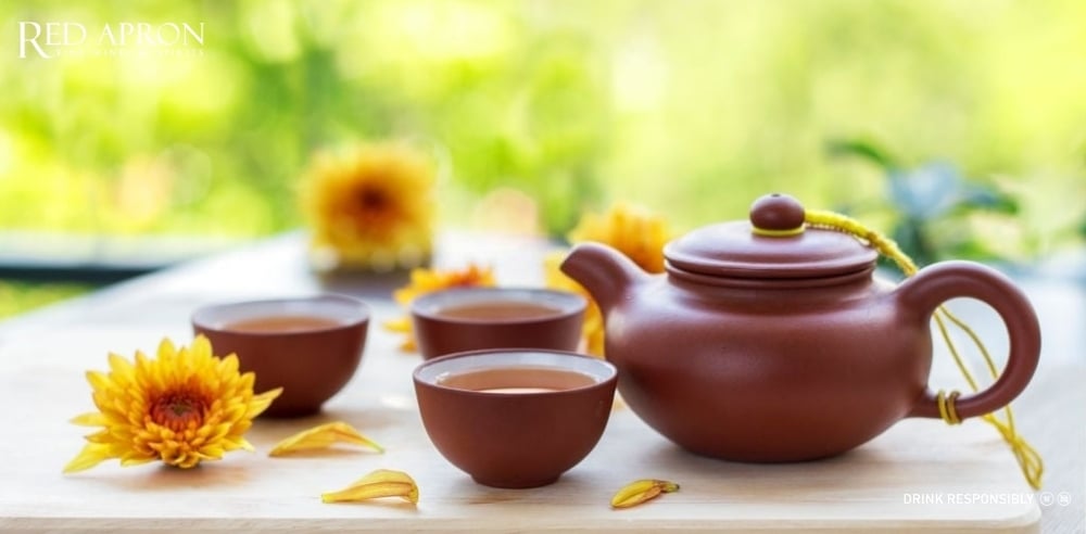 Bộ ấm trà và chén gốm màu nâu đỏ đặt cạnh hoa cúc vàng, minh họa ý tưởng quà Tết handmade là trà Tết/thảo mộc