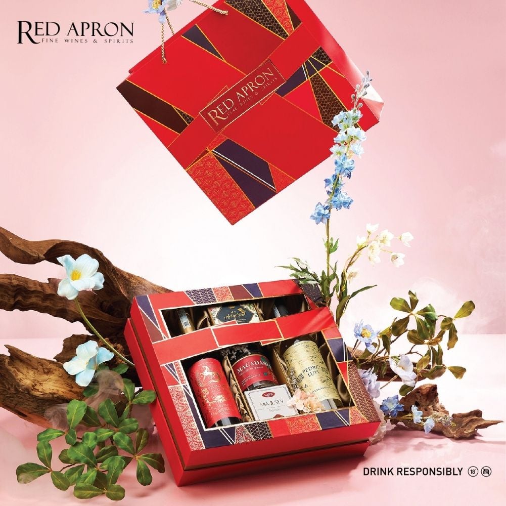 Ảnh hộp quà Tết màu đỏ in logo “RED APRON FINE WINES & SPIRITS”, nổi bật trên nền hồng, bên trong có nhiều chai rượu và hũ quà tặng, gợi ý địa chỉ mua quà tết tốt cho sức khỏe uy tín tại Việt Nam.