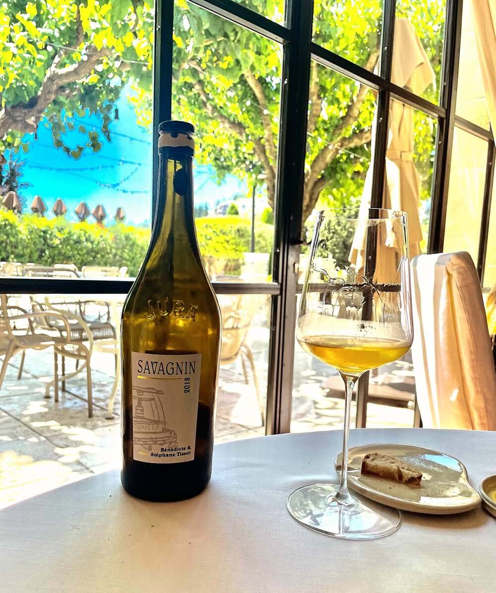 Chai Savagnin Bénédicte & Stéphane Tissot trên bàn cùng ly rượu vàng rơm, phong cách Jura.