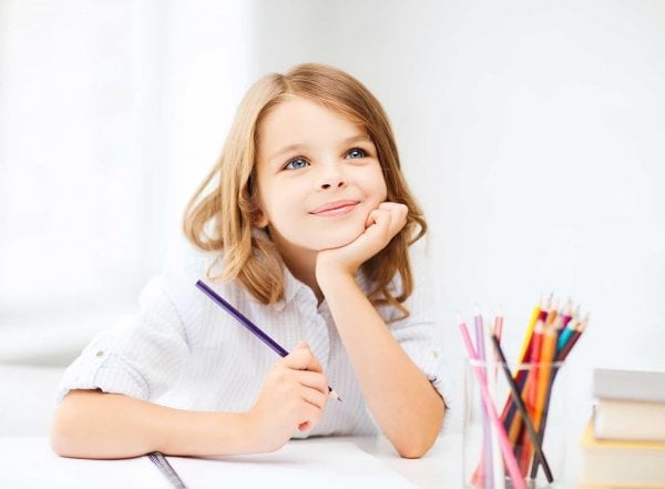 Bé gái chống cằm cầm bút chì bên chồng sách và hộp bút màu