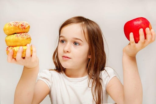 Bé gái so sánh giữa một tay cầm bánh ngọt và một tay cầm quả táo