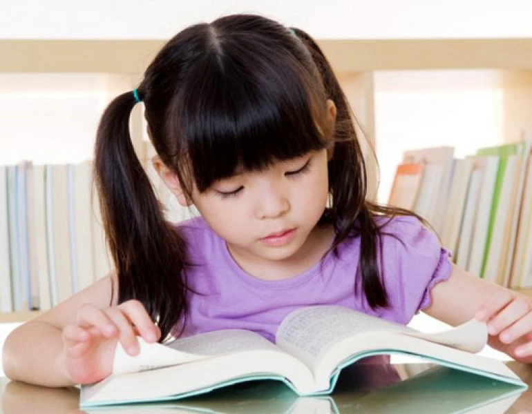 Bé gái ngồi đọc sách trước giá sách