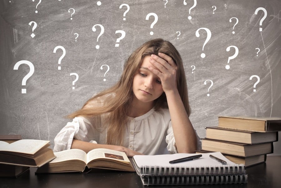 Nữ sinh ôm đầu trước bàn sách, phía sau là bảng đầy dấu hỏi