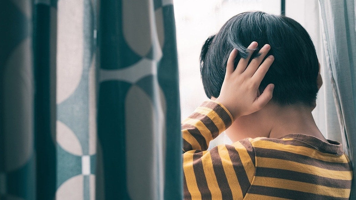 Bé trai đứng úp mặt vào cửa sổ, hai tay ôm sau đầu