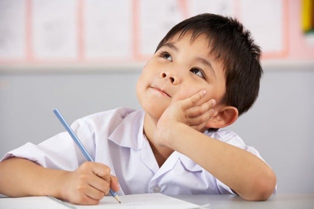 Bé trai mặc áo trắng chống tay lên má, nhìn xa khi đang làm bài
