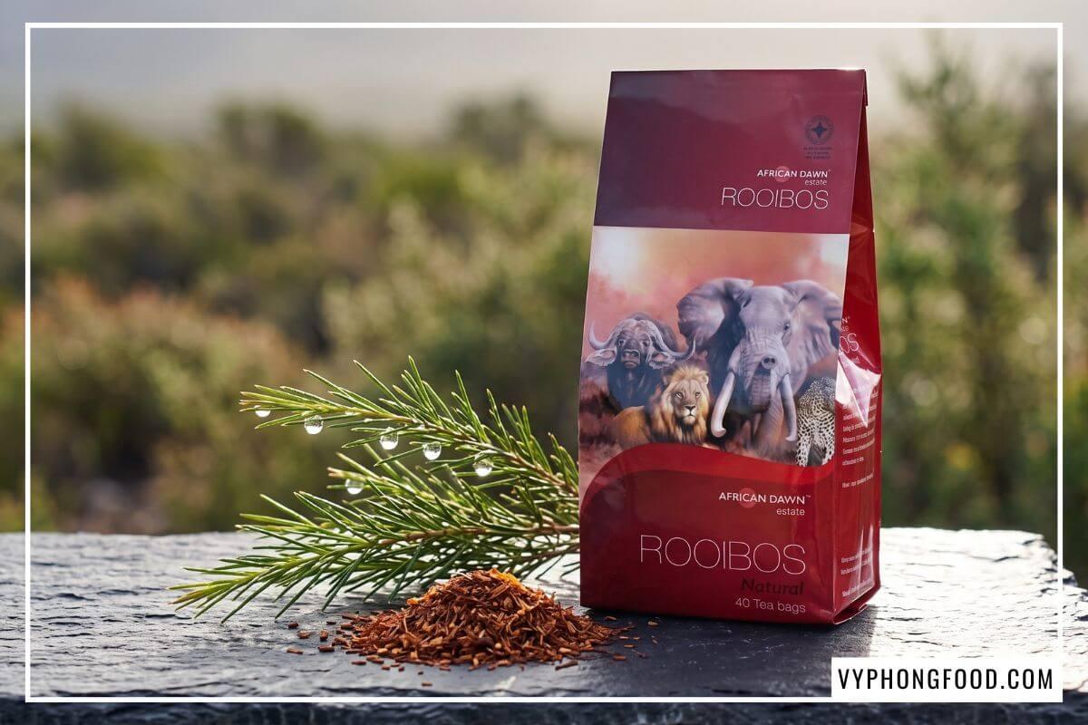 Gói trà không cafein màu đỏ đô đặt cạnh nhành lá kim tươi đọng sương trên nền đá phiến sần sùi.