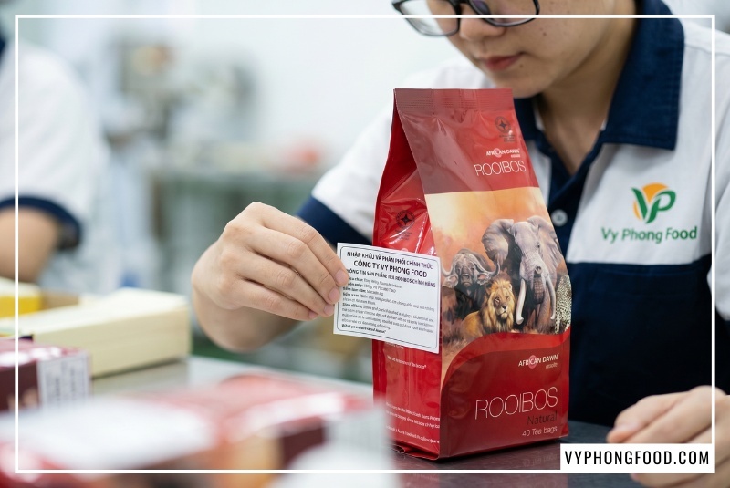Túi hồng trà rooibos nam phi African Dawn nhập khẩu chính ngạch đặt cạnh tách hồng trà rooibos nam phi màu đỏ hổ phách. Giải pháp hoàn hảo cho câu hỏi trà rooibos mua ở đâu để đảm bảo an toàn, không nấm mốc và tận dụng tối đa công dụng của trà thảo mộc tốt cho sức khỏe gia đình.