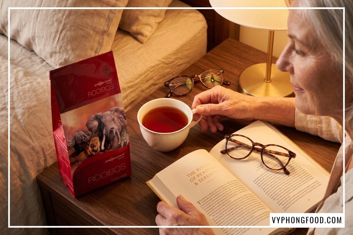 Cụ bà đọc sách bên tách trà rooibos túi lọc cho người cao tuổi và gói trà đỏ African Dawn trên bàn gỗ.