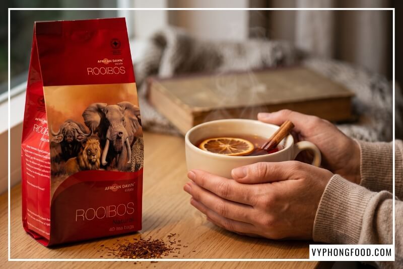 Tách trà thảo mộc thiên nhiên đỏ hổ phách ấm áp bên gói rooibos African Dawn góc bàn gỗ sồi.